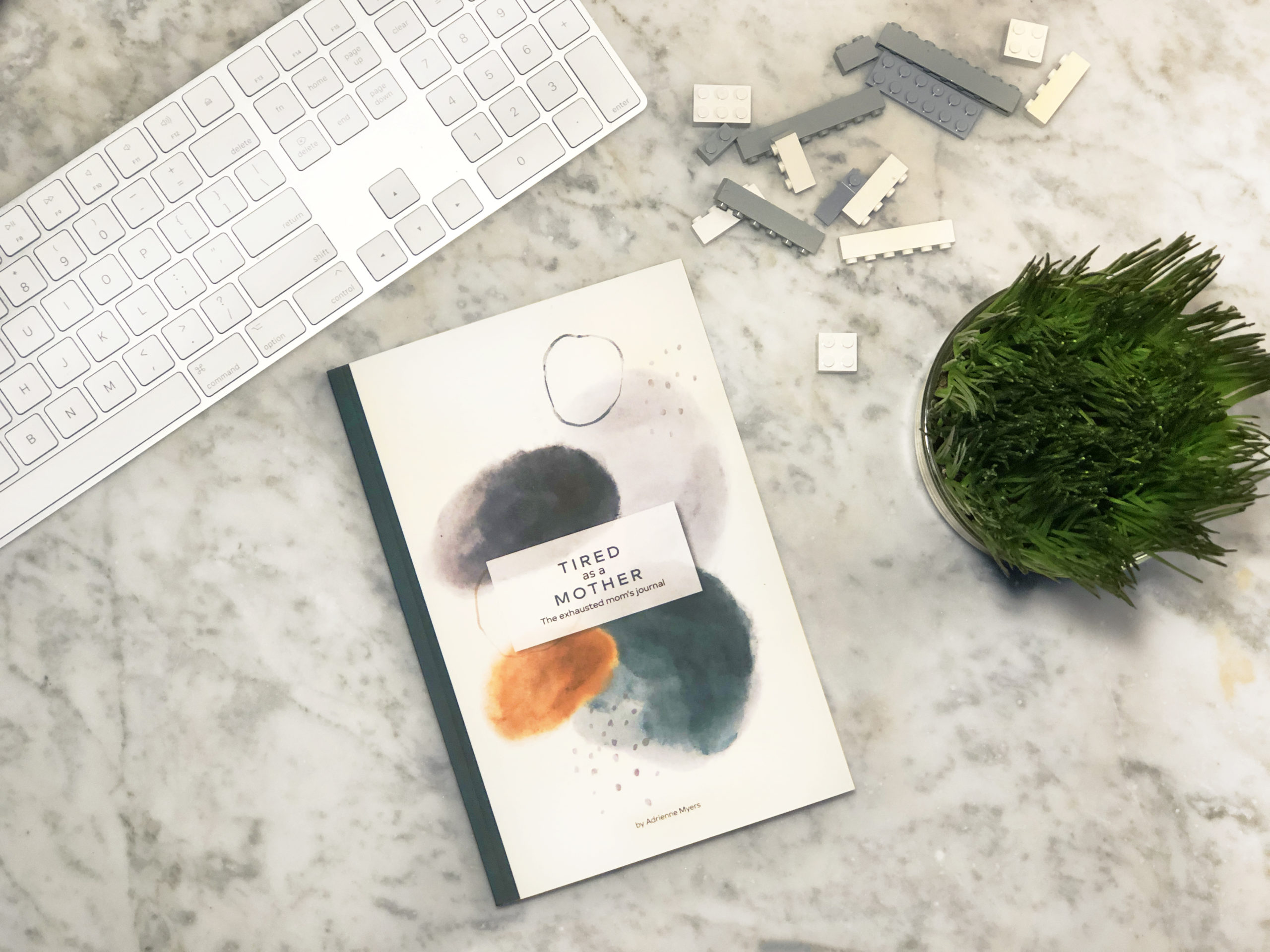 Tired as a mother: the exhausted mom's journal book on a white and grey marble background with keyboard, legos and plant